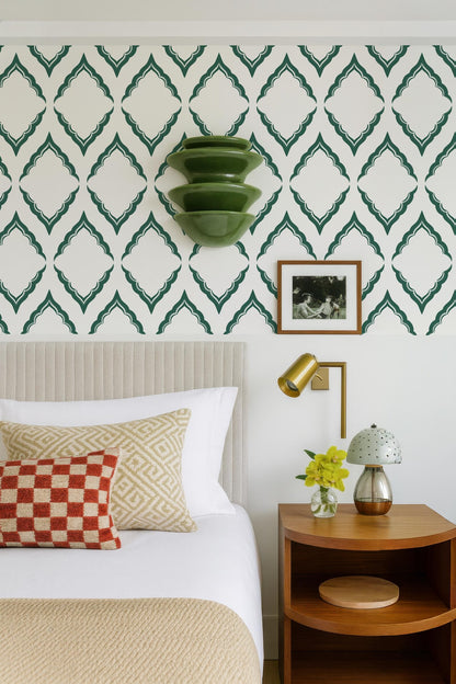a well-decorated bedroom with a bed, nightstand, and wall art. The bed has a white comforter and a red and white checkered pillow. The nightstand has a lamp and a vase with yellow flowers. The wall behind the bed has a patterned wallpaper with green diamond shapes. There is a framed photo hanging on the wall, and a green wall hanging with three stacked bowls.