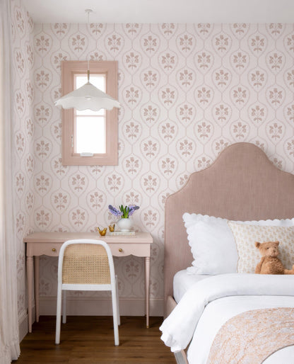 a cozy bedroom with a pink and white floral wallpaper, a beige headboard, and a white chair. A teddy bear is sitting on the bed, and there is a vase of flowers on the desk.