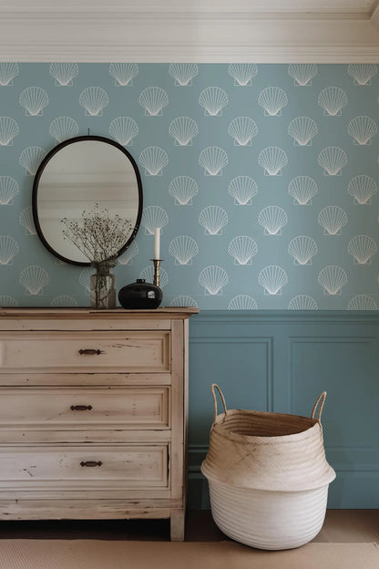 a light-colored wooden dresser with a round mirror above it, set against a blue wall with white shell patterns. The dresser also has a small vase of flowers and a candle on top. Next to the dresser is a large woven basket.