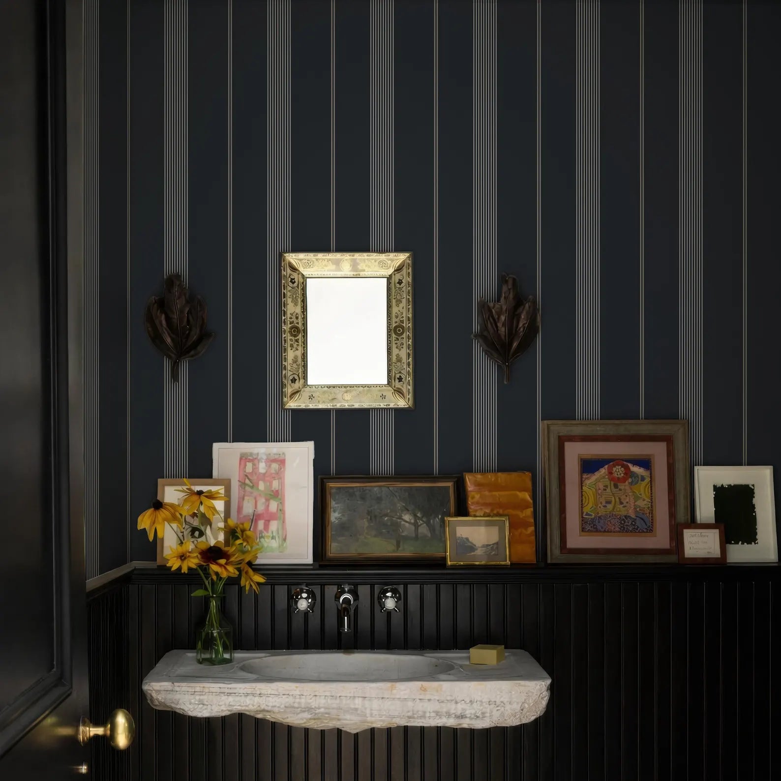 a bathroom with a black and white striped wall, a white sink, and a shelf with framed pictures and a vase of yellow flowers.