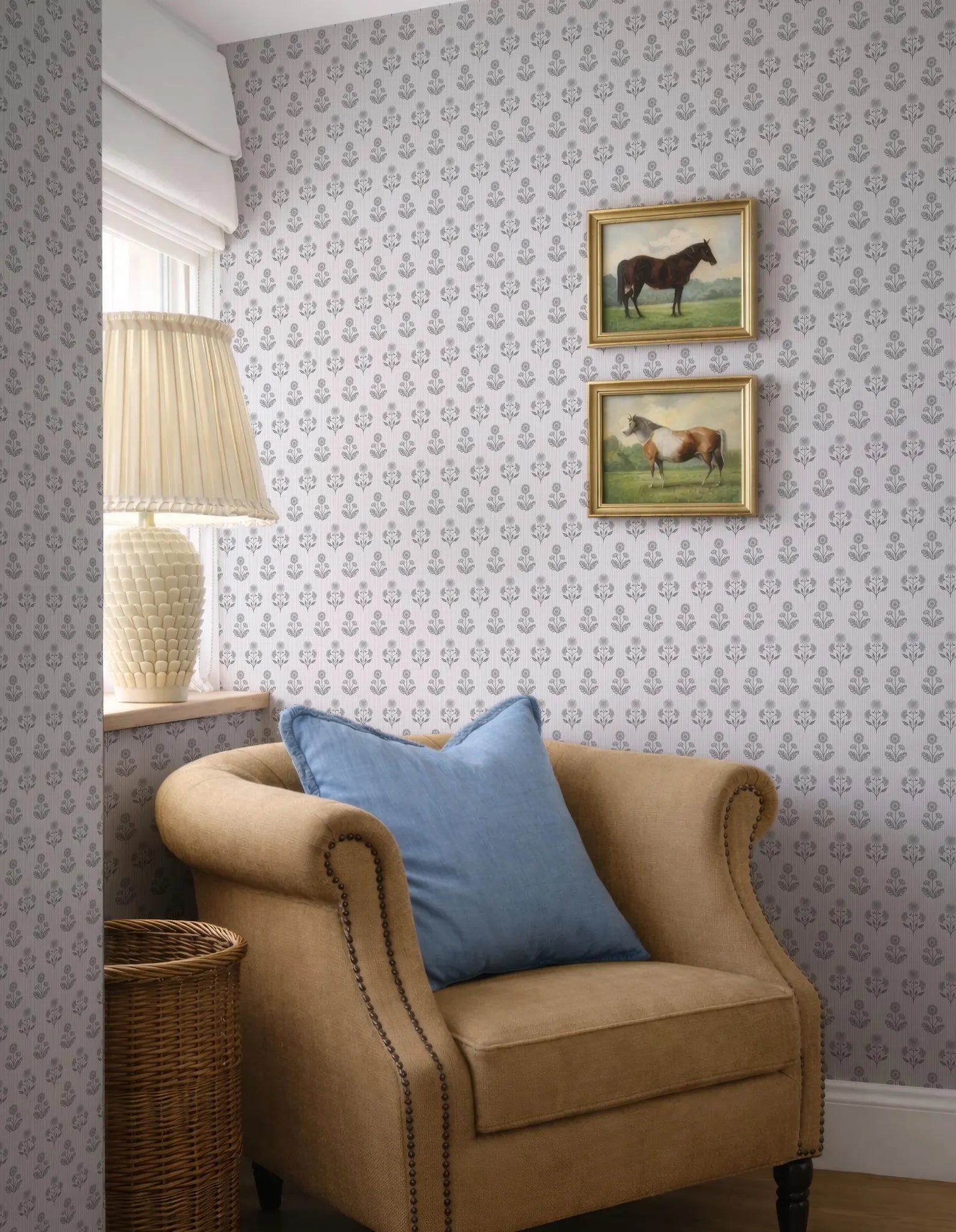 a cozy corner of a room with a beige armchair, a wicker basket, and a lamp. Above the armchair, there are two framed pictures of horses, adding a touch of nature and elegance to the space. The wall behind the armchair is adorned with a patterned wallpaper, complementing the overall design of the room.