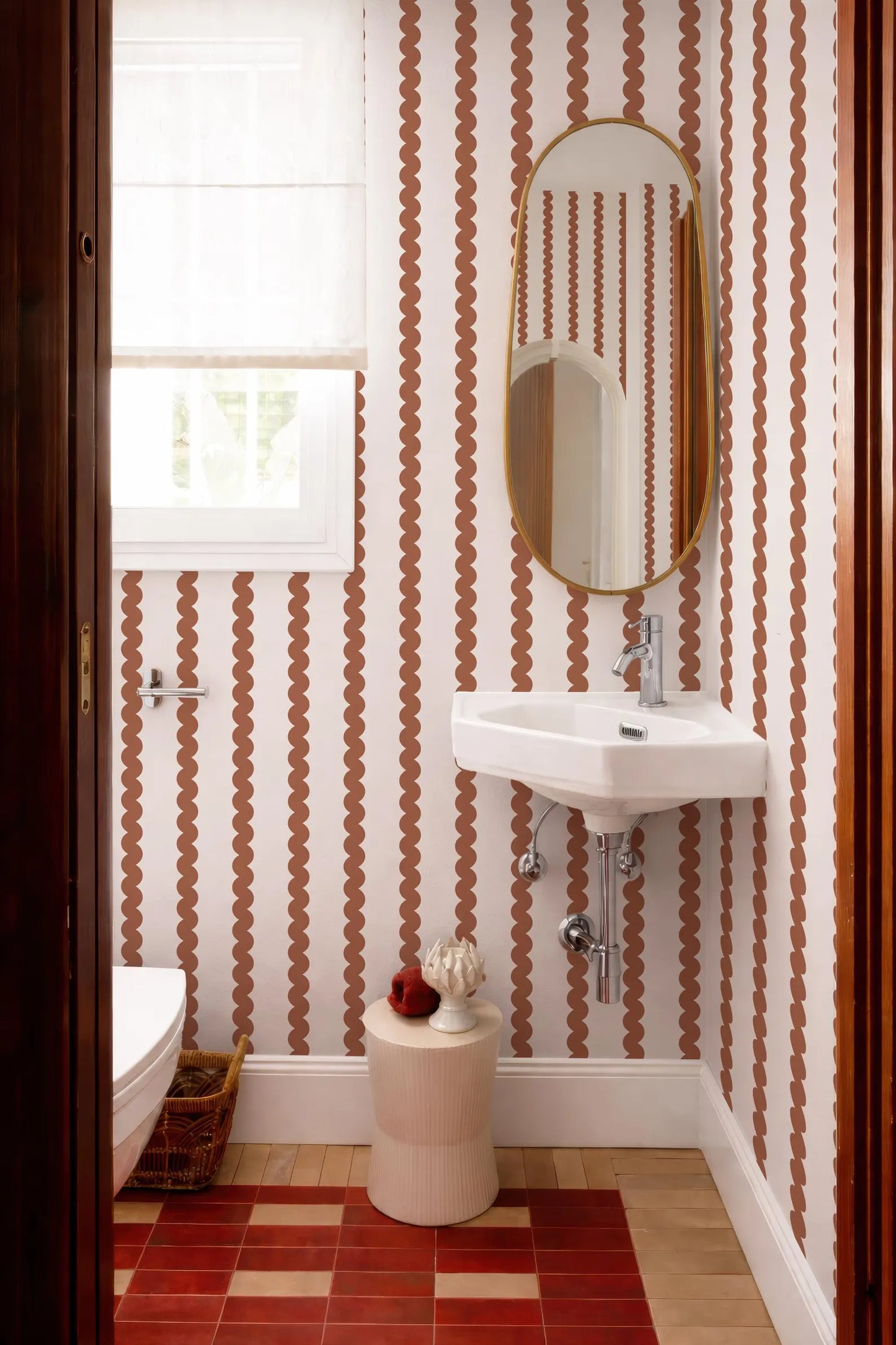 a bathroom with a white sink, a mirror, and a window. The walls are covered in a patterned wallpaper with a repeating design of orange and white circles. The floor is made of red tiles.