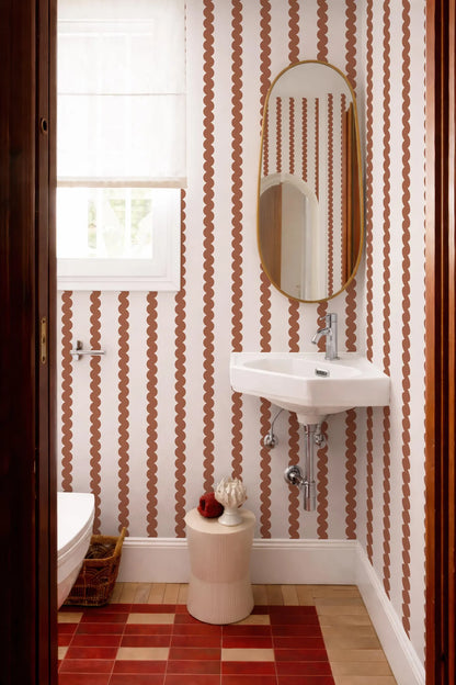 a bathroom with a white sink, a mirror, and a window. The walls are covered in a patterned wallpaper with a repeating design of orange and white circles. The floor is made of red tiles.