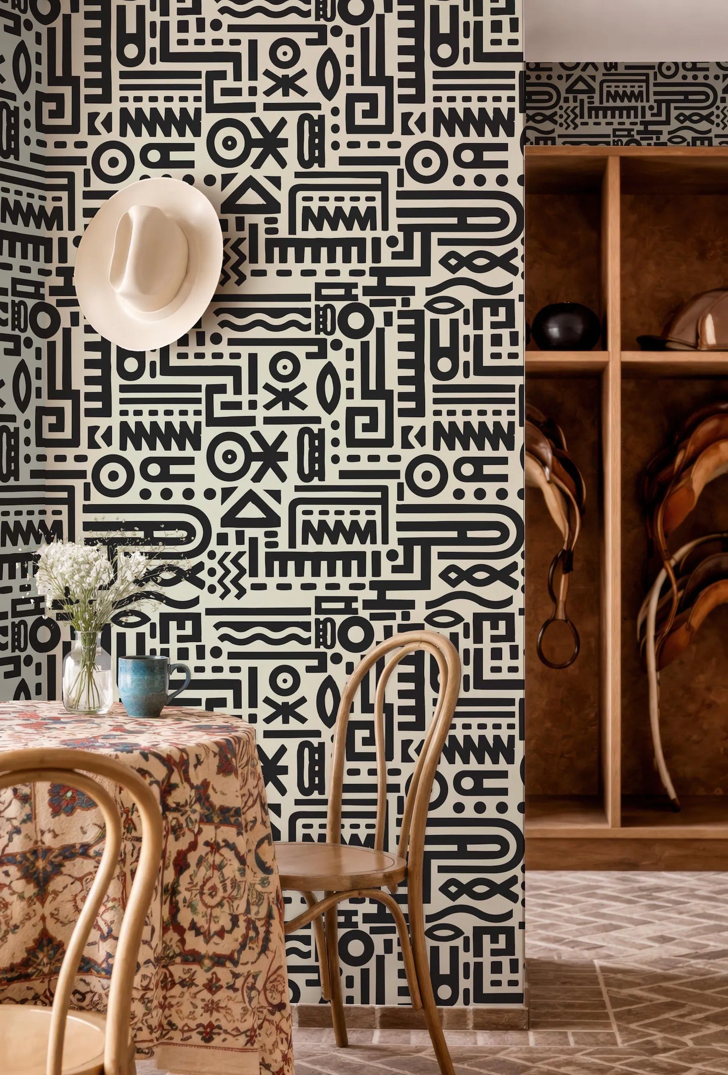 a room with a black and white patterned wall, a table with a floral tablecloth, and a wooden shelf with various items.