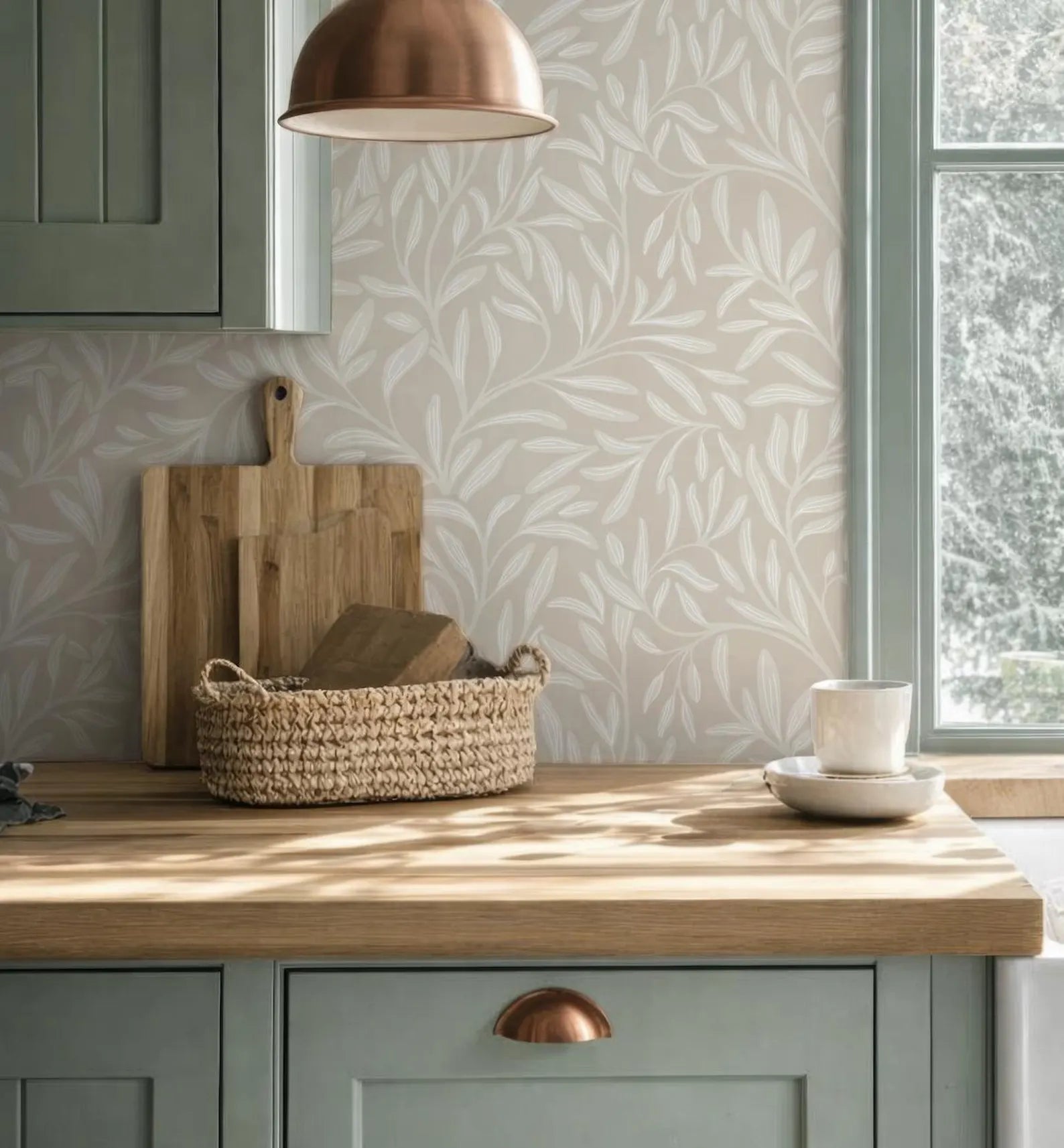 a kitchen counter with a woven basket, a wooden cutting board, and a white coffee cup on a light green cabinet. The wall behind the counter has a patterned wallpaper, and a copper pendant light hangs above the counter.