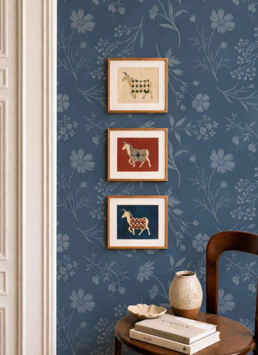 a cozy corner of a room with a blue floral wallpaper, a wooden chair, and a small table with a vase and a book. There are three framed pictures hanging on the wall, depicting animals in different colors.