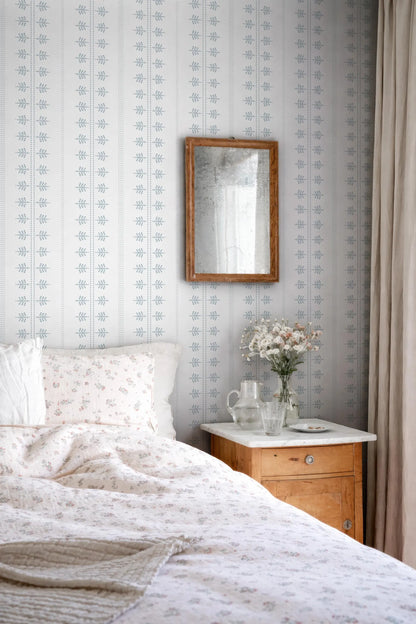 a cozy bedroom scene with a bed, a wooden nightstand, and a mirror on the wall. The bed is covered in a white and floral comforter, and there is a vase of flowers on the nightstand. The mirror is hung above the nightstand, which also has a small table and a lamp. The walls are decorated with a patterned wallpaper, and there is a window with curtains in the background.