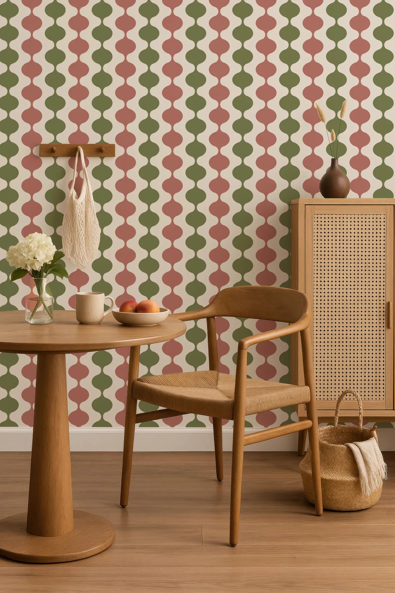a cozy dining area with a wooden table, chair, and woven cabinet. The wall behind the table has a repeating pattern of green and red circles, creating a vibrant and lively atmosphere. On the table, there is a bowl of fruit, a vase with flowers, and a cup. The overall scene conveys a warm and inviting ambiance.