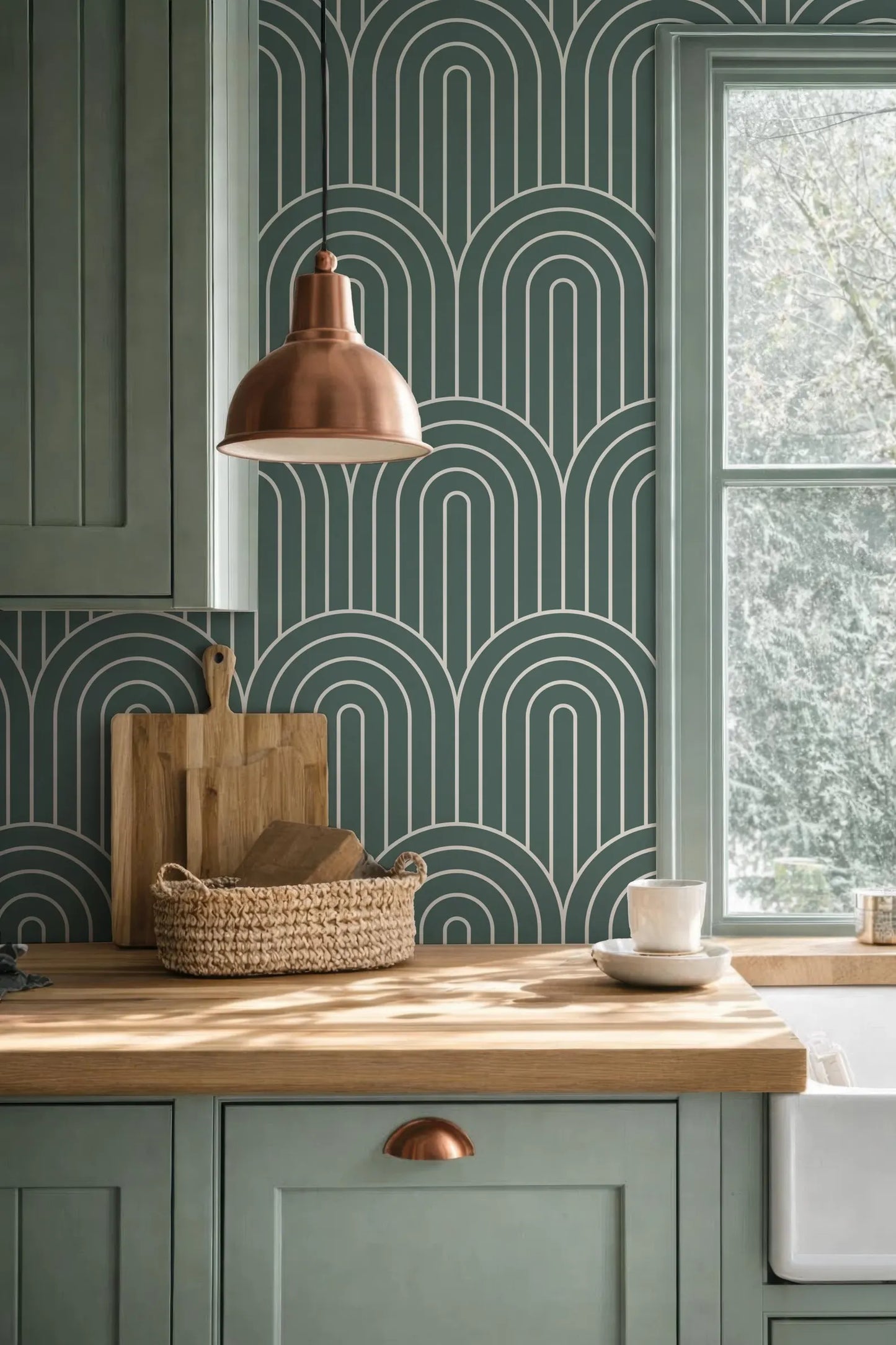 a kitchen with a wooden countertop, a woven basket, a wooden cutting board, and a copper pendant light. The kitchen has green cabinets and a patterned wallpaper, creating a cohesive and stylish design.