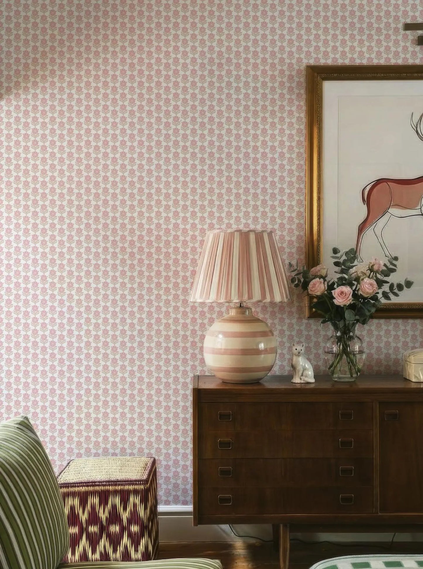a cozy living room with a pink and white patterned wallpaper, a wooden dresser with a lamp and a vase of flowers, and a striped ottoman.