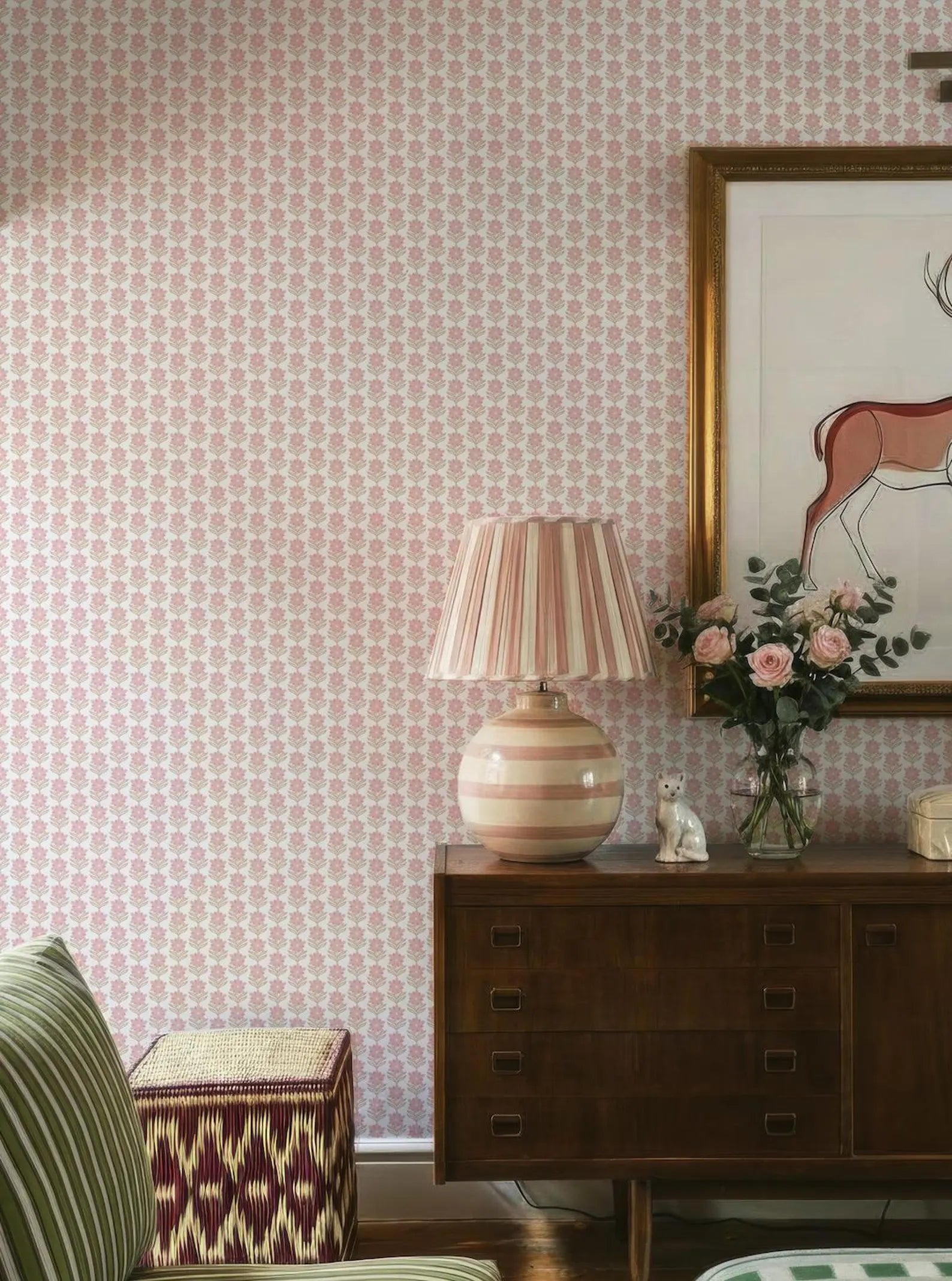 a cozy living room with a pink and white patterned wallpaper, a wooden dresser with a lamp and a vase of flowers, and a striped ottoman.