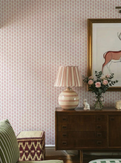 a cozy living room with a pink and white patterned wallpaper, a wooden dresser with a lamp and a vase of flowers, and a striped ottoman.