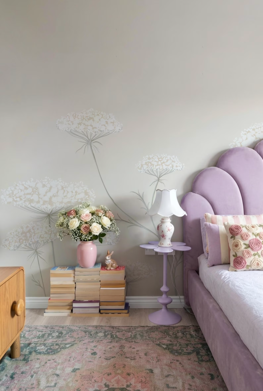 a bedroom with a purple headboard, a nightstand with a lamp and a vase of flowers, and a stack of books on the floor.