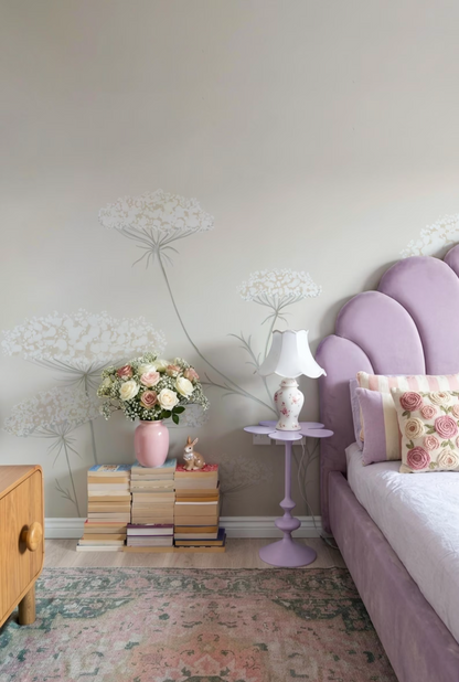 a bedroom with a purple headboard, a nightstand with a lamp and a vase of flowers, and a stack of books on the floor.