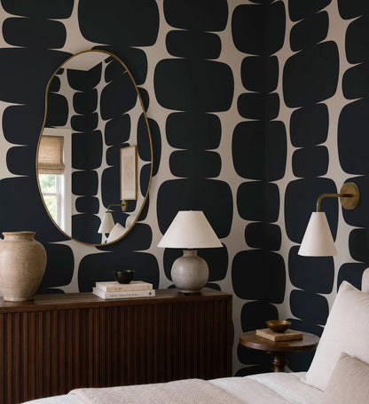 a bedroom with a large mirror, a wooden dresser, and a lamp on a side table. The walls are covered in a bold black and white geometric pattern, creating a striking visual effect.