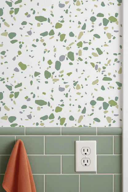 a bathroom wall with a pattern of green and gray tiles, and a white electrical outlet is mounted on the wall above the tiles. There is also a red towel hanging on the wall.