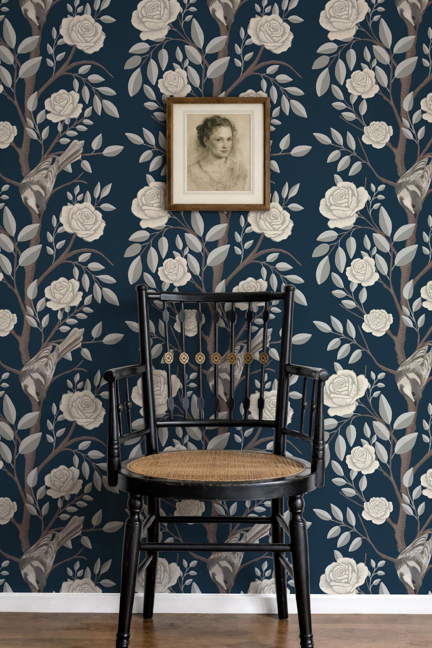 a black wooden chair with a woven seat, positioned in front of a wall with a floral wallpaper pattern. Above the chair, there is a framed portrait of a woman.