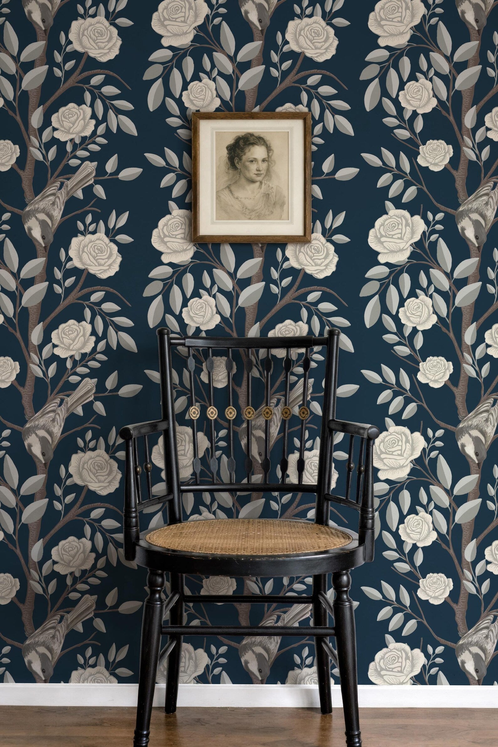 a black wooden chair with a woven seat, positioned in front of a wall with a floral wallpaper pattern. Above the chair, there is a framed portrait of a woman.