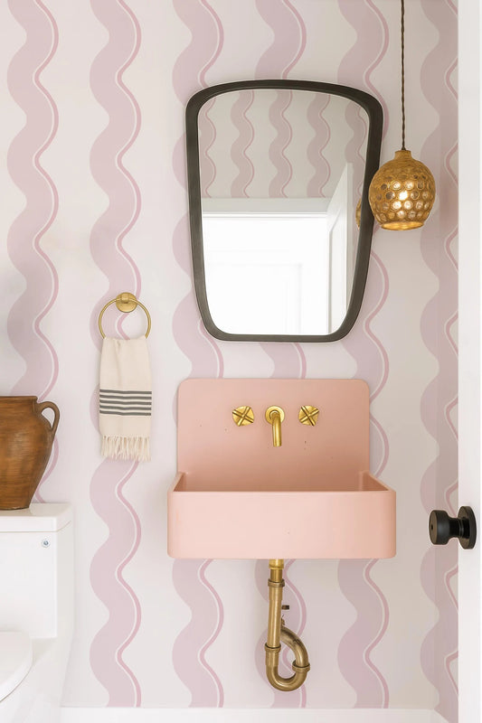 a bathroom with a pink sink, a black mirror, and a gold pendant light. The walls are decorated with pink and white stripes, and there is a wooden vase on the counter.