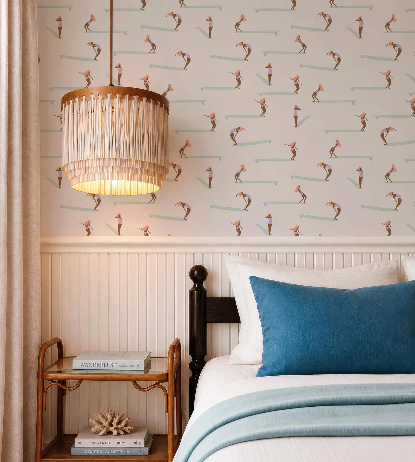 a bedroom with a bed, a nightstand, and a chandelier. The bed has a blue pillow and a white comforter. The nightstand has a lamp and a book. The wall behind the bed has a pattern of surfers on a wave.