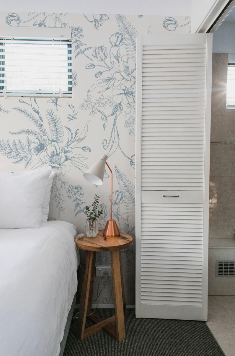 a cozy bedroom scene with a bed, a nightstand, and a window with blinds. The room has a floral wallpaper and a white wooden door with vertical slats.