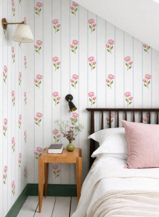 a cozy bedroom with a bed, a wooden nightstand, and a lamp. The walls are covered in a floral wallpaper with pink flowers, and the room has a white color scheme.