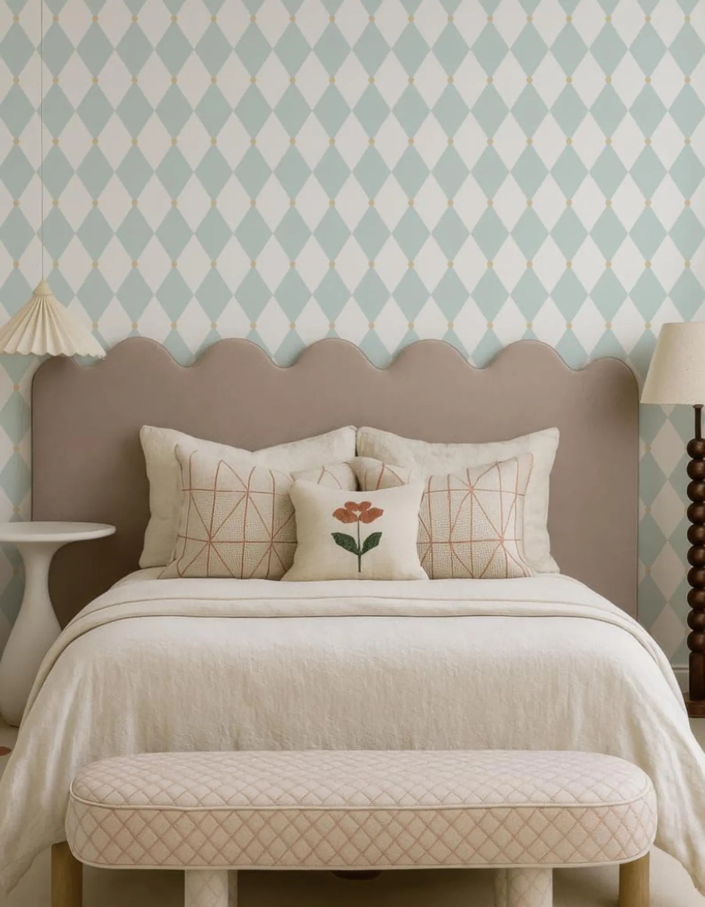 a cozy bedroom with a bed, a nightstand, and a lamp. The bed has a gray headboard and is adorned with a white comforter and a single red flower pillow. The nightstand has a white base and a lamp with a beige shade. The walls are covered in a diamond patterned wallpaper, and the overall color scheme is predominantly white and beige.