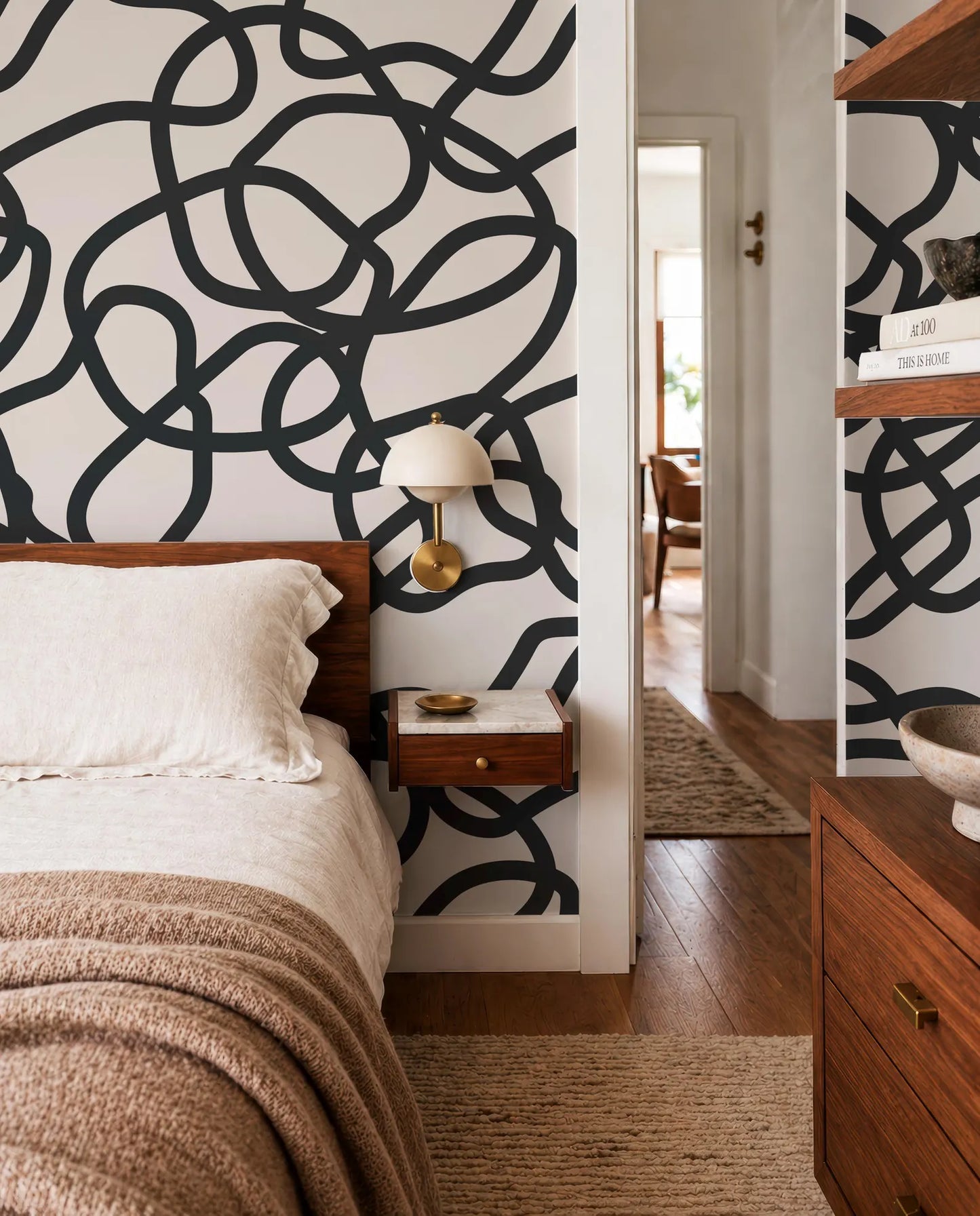 a modern bedroom with a large black and white abstract design on the wall behind the bed. The room has a wooden dresser and a nightstand with a lamp, creating a cozy and stylish atmosphere.