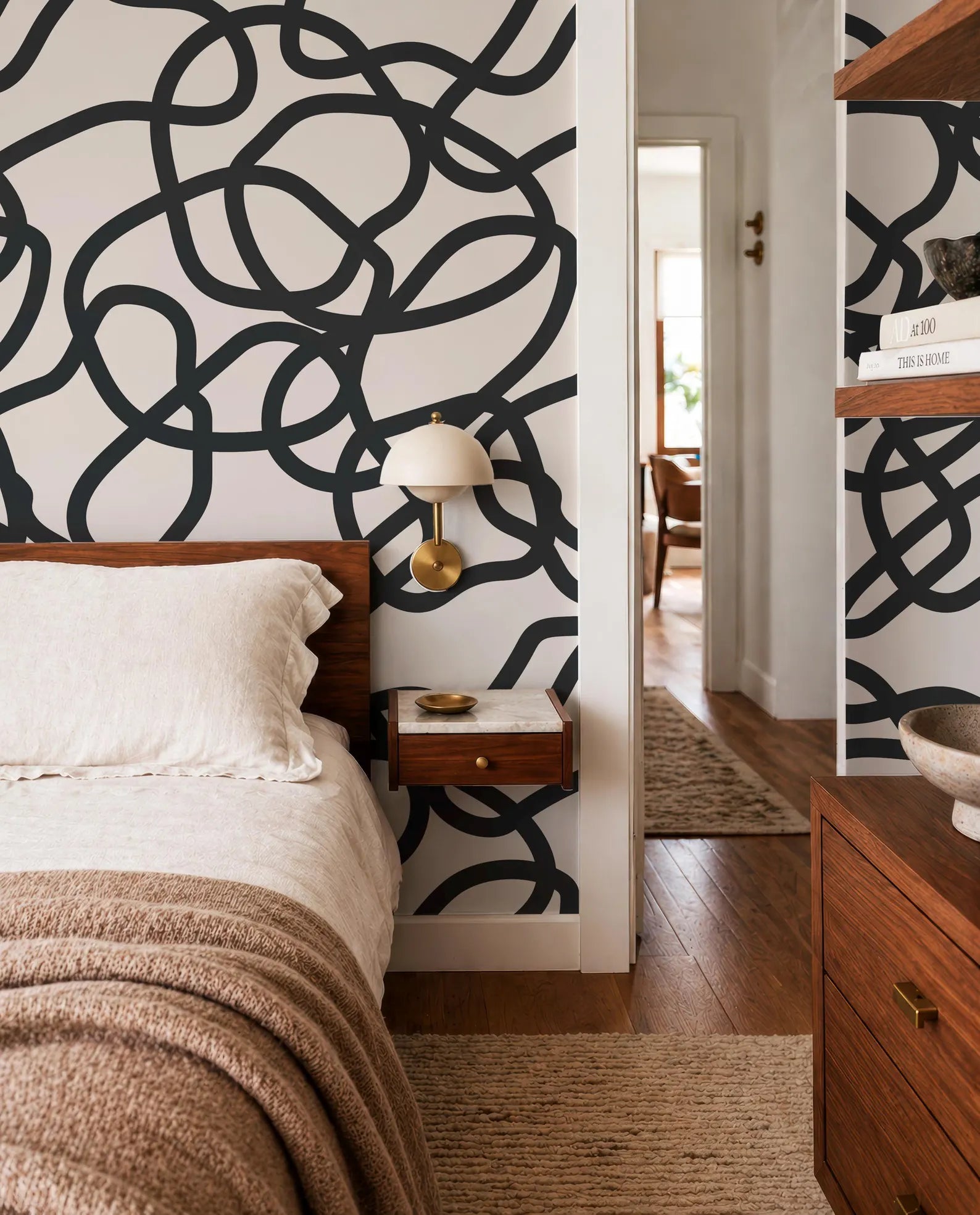 a modern bedroom with a large black and white abstract design on the wall behind the bed. The room has a wooden dresser and a nightstand with a lamp, creating a cozy and stylish atmosphere.