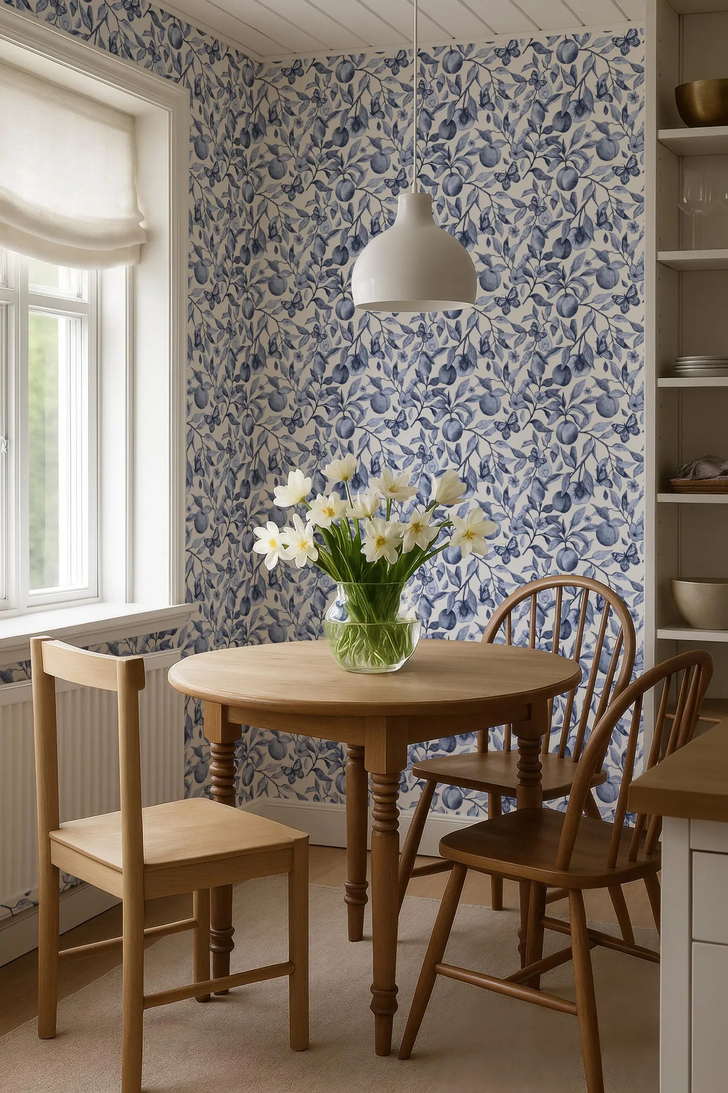 a cozy dining area with a round wooden table, two wooden chairs, and a vase of white flowers on the table. The walls are covered in blue and white floral wallpaper, and a white pendant light hangs above the table.
