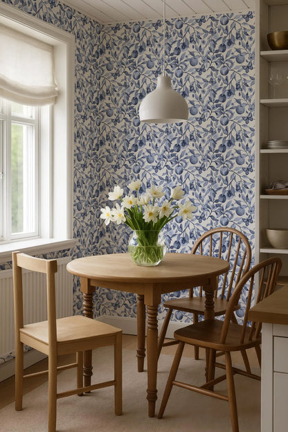 a cozy dining area with a round wooden table, two wooden chairs, and a vase of white flowers on the table. The walls are covered in blue and white floral wallpaper, and a white pendant light hangs above the table.
