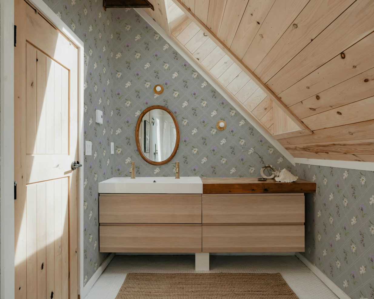 a bathroom with a wooden vanity and a mirror above it. The walls are covered in a floral patterned wallpaper, and the ceiling has a slanted design.