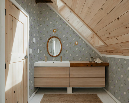 a bathroom with a wooden vanity and a mirror above it. The walls are covered in a floral patterned wallpaper, and the ceiling has a slanted design.