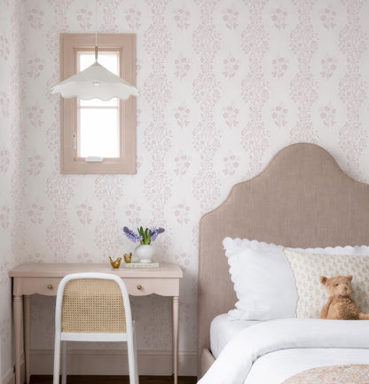 a cozy bedroom with a bed, a desk, and a window. The bed has a beige headboard and white bedding, and a teddy bear is resting on it. The desk has a white chair and a vase of purple flowers, and there is a small gold bird figurine on the desk. The wall behind the desk has a floral wallpaper pattern, and there is a window with a white shade.