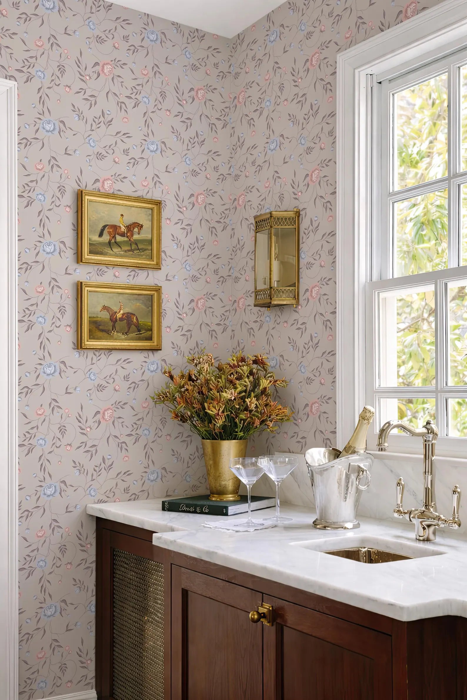 a cozy corner of a room with a white countertop, a vase of flowers, and a set of wine glasses. The wall behind the countertop has a floral wallpaper pattern, and there are two framed pictures hanging on the wall. A window is visible on the right side of the image, allowing natural light to enter the space.