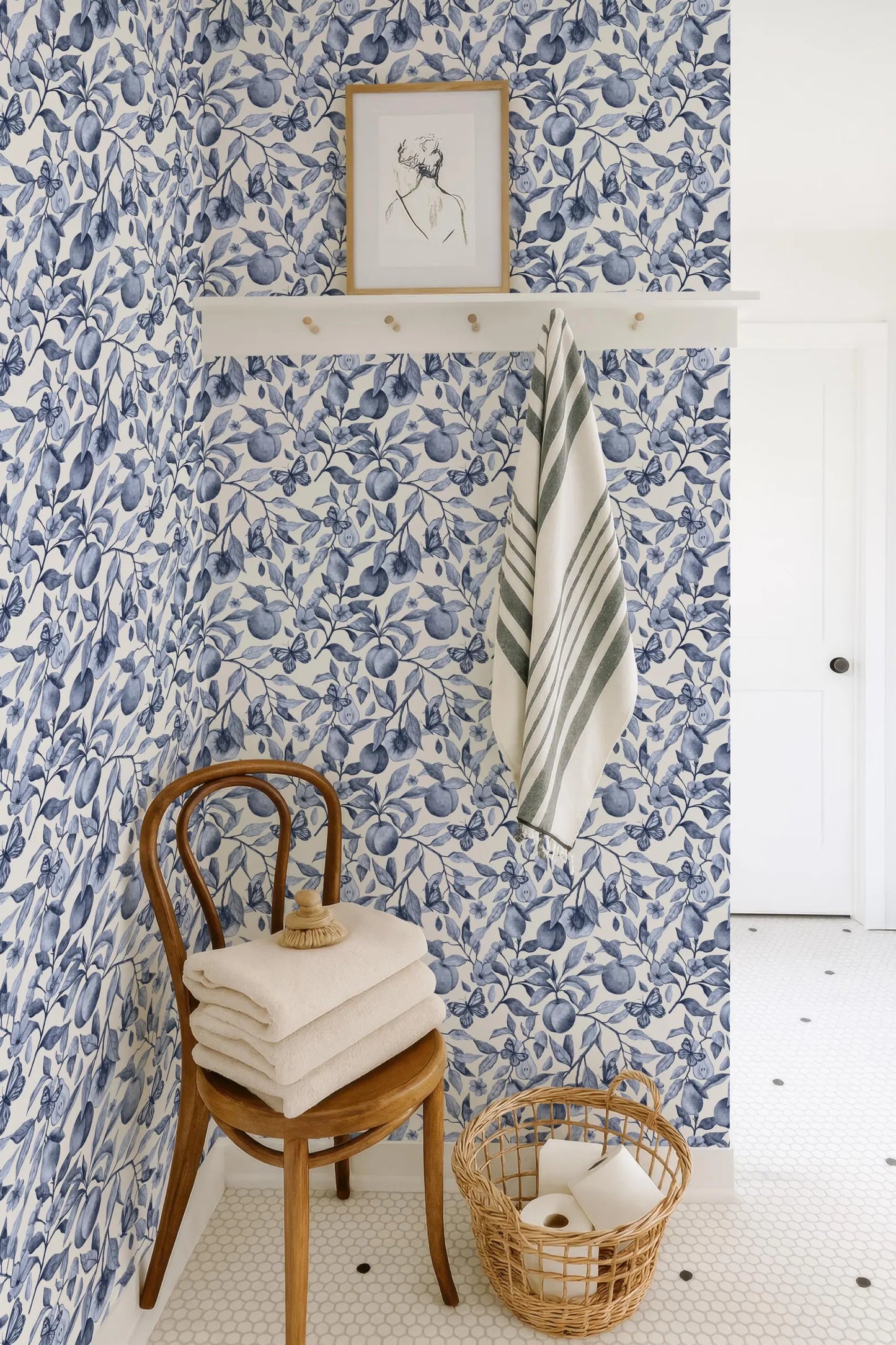 a bathroom with blue and white floral wallpaper, a wooden chair with folded towels, a basket of toilet paper, and a framed picture on the wall.