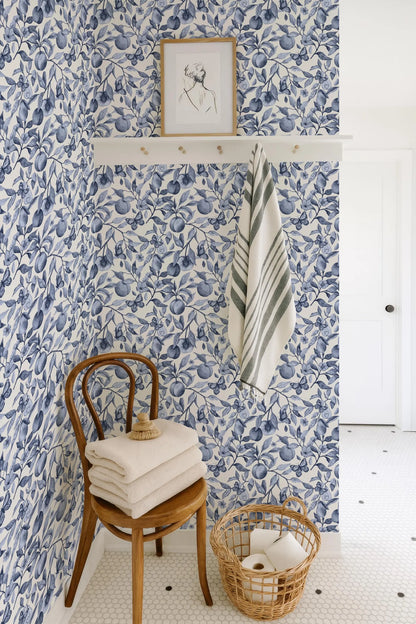 a bathroom with blue and white floral wallpaper, a wooden chair with folded towels, a basket of toilet paper, and a framed picture on the wall.