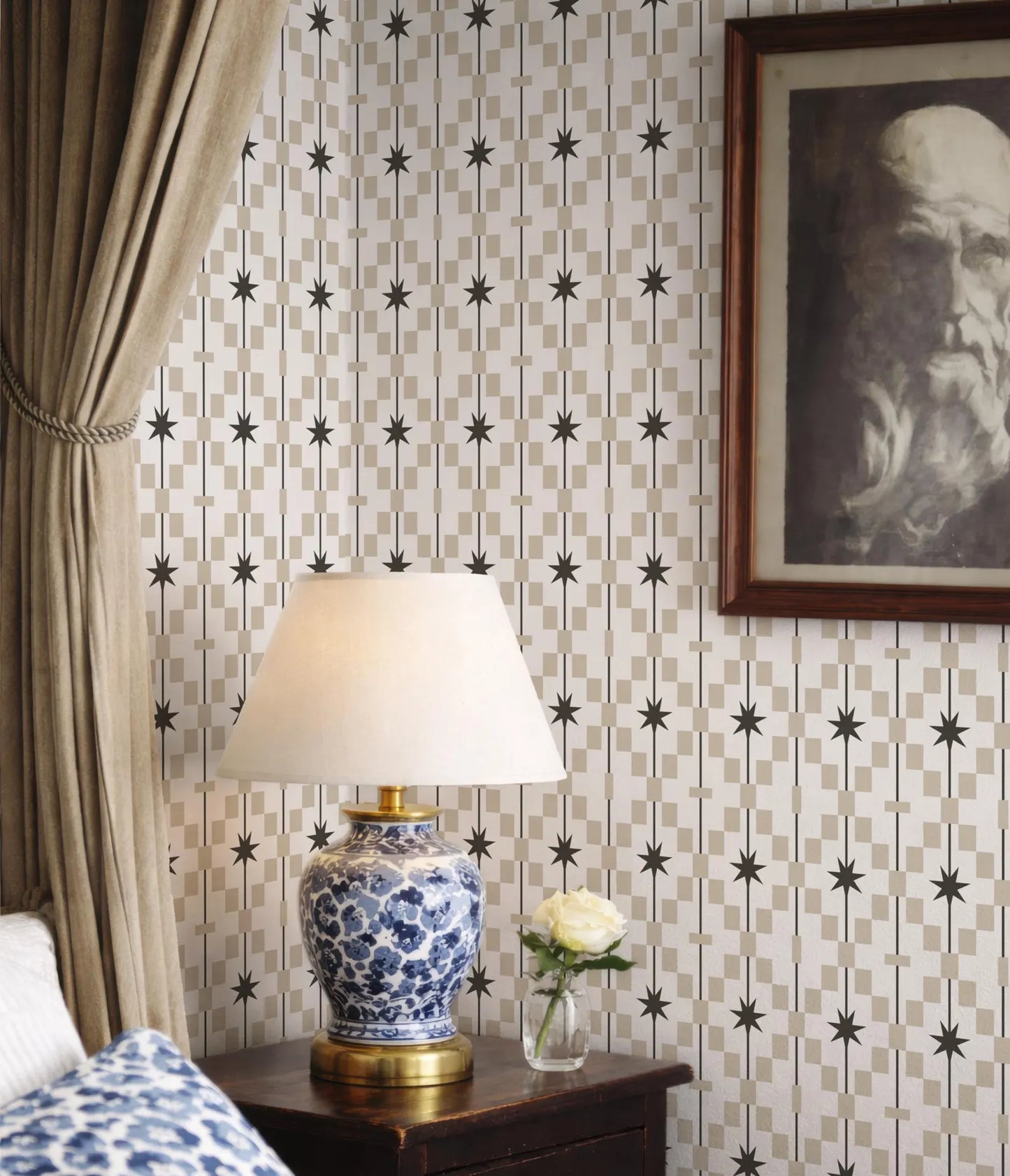 a bedroom setting with a blue and white patterned lamp on a wooden nightstand, a framed portrait of a man on the wall, and a white rose in a vase.
