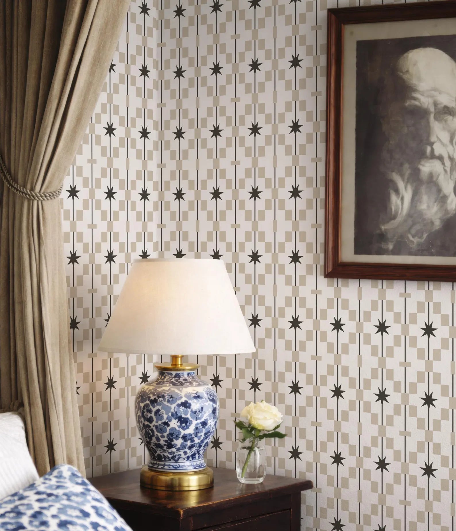 a bedroom setting with a blue and white patterned lamp on a wooden nightstand, a framed portrait of a man on the wall, and a white rose in a vase.