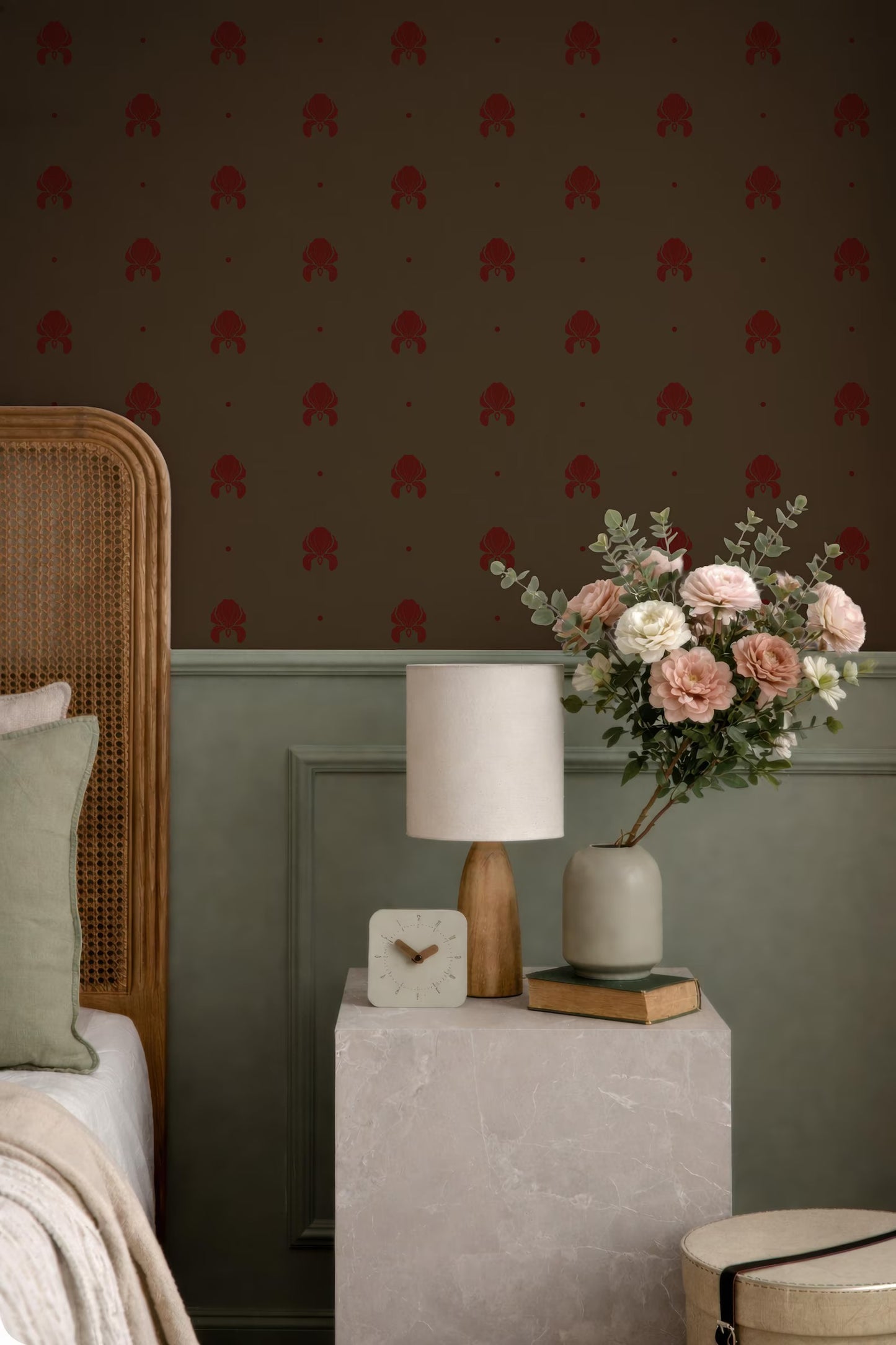 a bedroom setting with a wooden headboard, a white lamp, a vase of flowers, and a small table with a clock and a book.