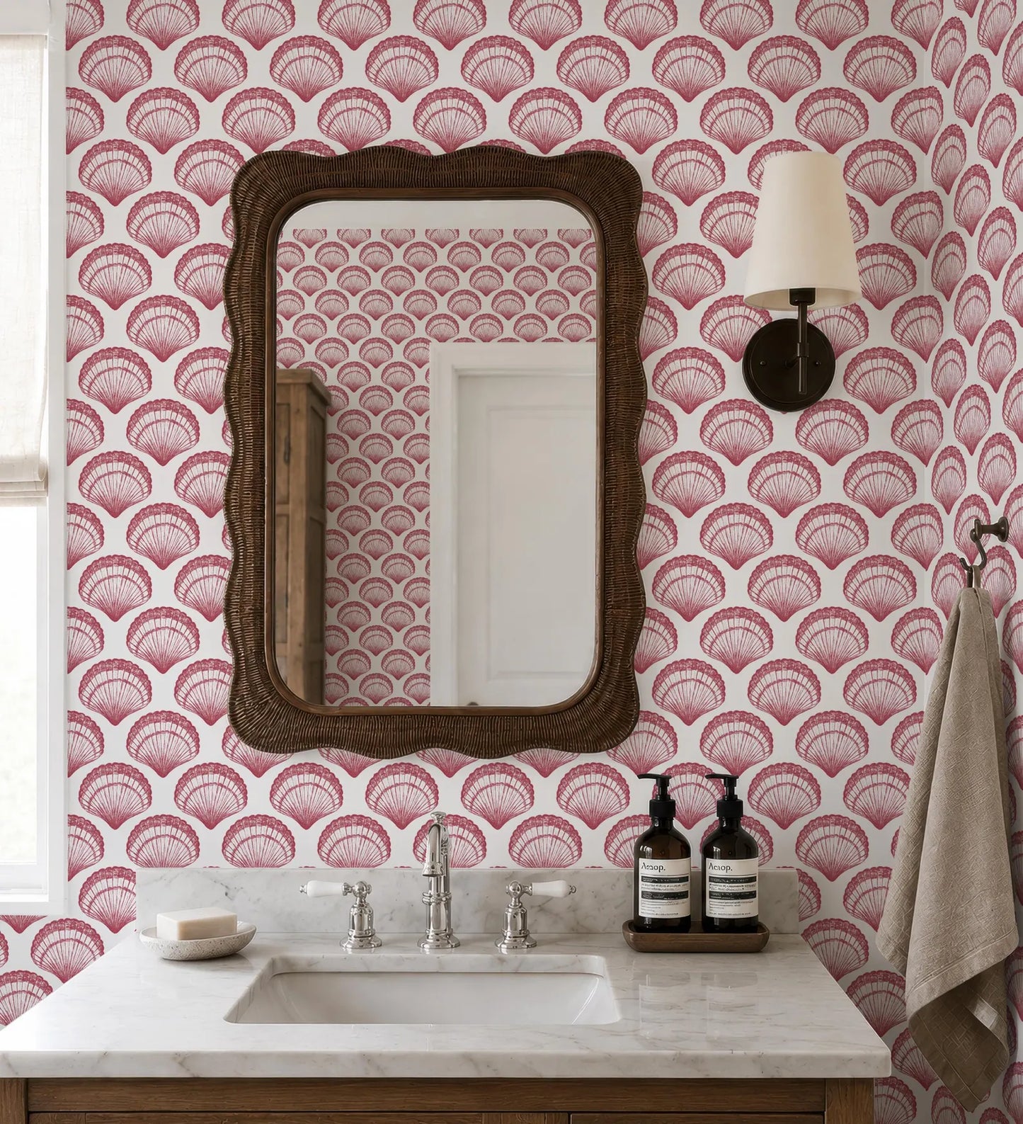 a bathroom with a white sink, a mirror, and a lamp. The walls are covered in a pink and white patterned wallpaper, and there are two bottles of soap on the counter.