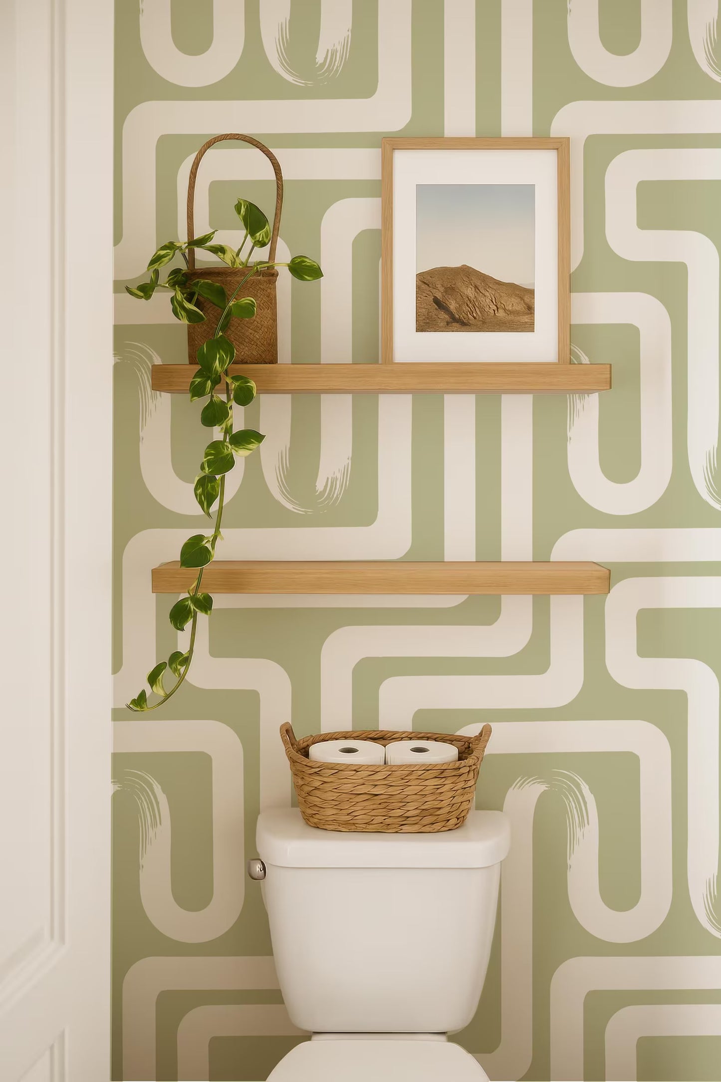 a bathroom with a white toilet, a wooden shelf above it, and a basket of toilet paper on the tank. The wall behind the toilet has a green and white patterned wallpaper.