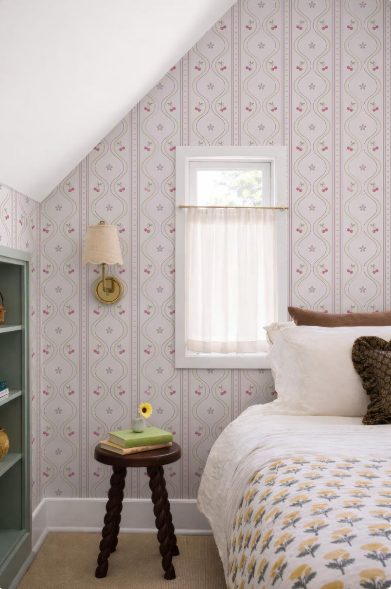 a cozy bedroom with a bed, a small wooden stool, and a window with a white curtain. The walls are covered in a patterned wallpaper, and there is a lamp on the wall. The room has a warm and inviting atmosphere.