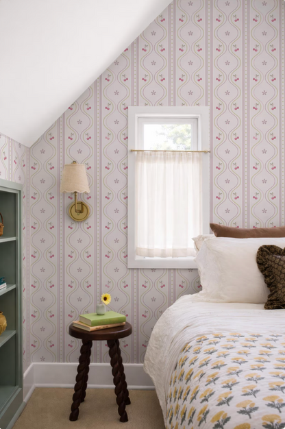 a cozy bedroom with a bed, a small wooden stool, and a window with a white curtain. The walls are covered in a patterned wallpaper, and there is a lamp on the wall. The room has a warm and inviting atmosphere.