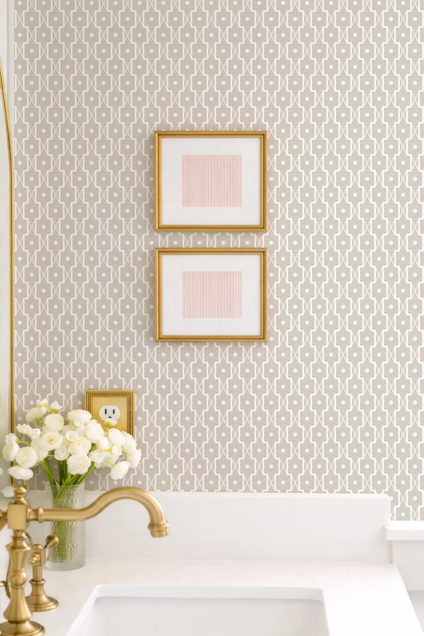 a bathroom with a white sink, a gold faucet, and a framed picture on the wall above the sink. There is also a vase of white flowers on the counter.