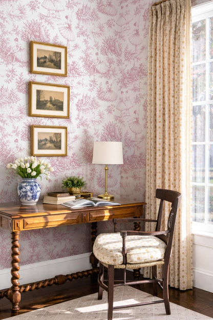 a cozy corner of a room with a wooden desk, a lamp, and a chair. The walls are adorned with pink floral wallpaper, and there are three framed pictures hanging above the desk. A vase of flowers and a book are also present on the desk, creating a warm and inviting atmosphere.
