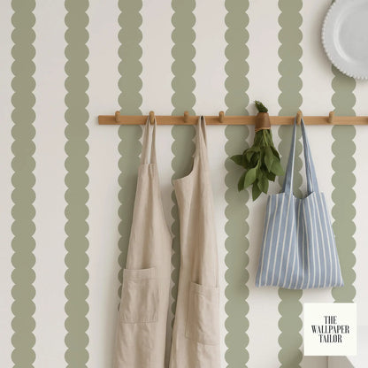 a wall with a wooden rack holding two white aprons and a striped blue and white bag. The wall has a pattern of green and white circles, and there is a small plant hanging from the rack.
