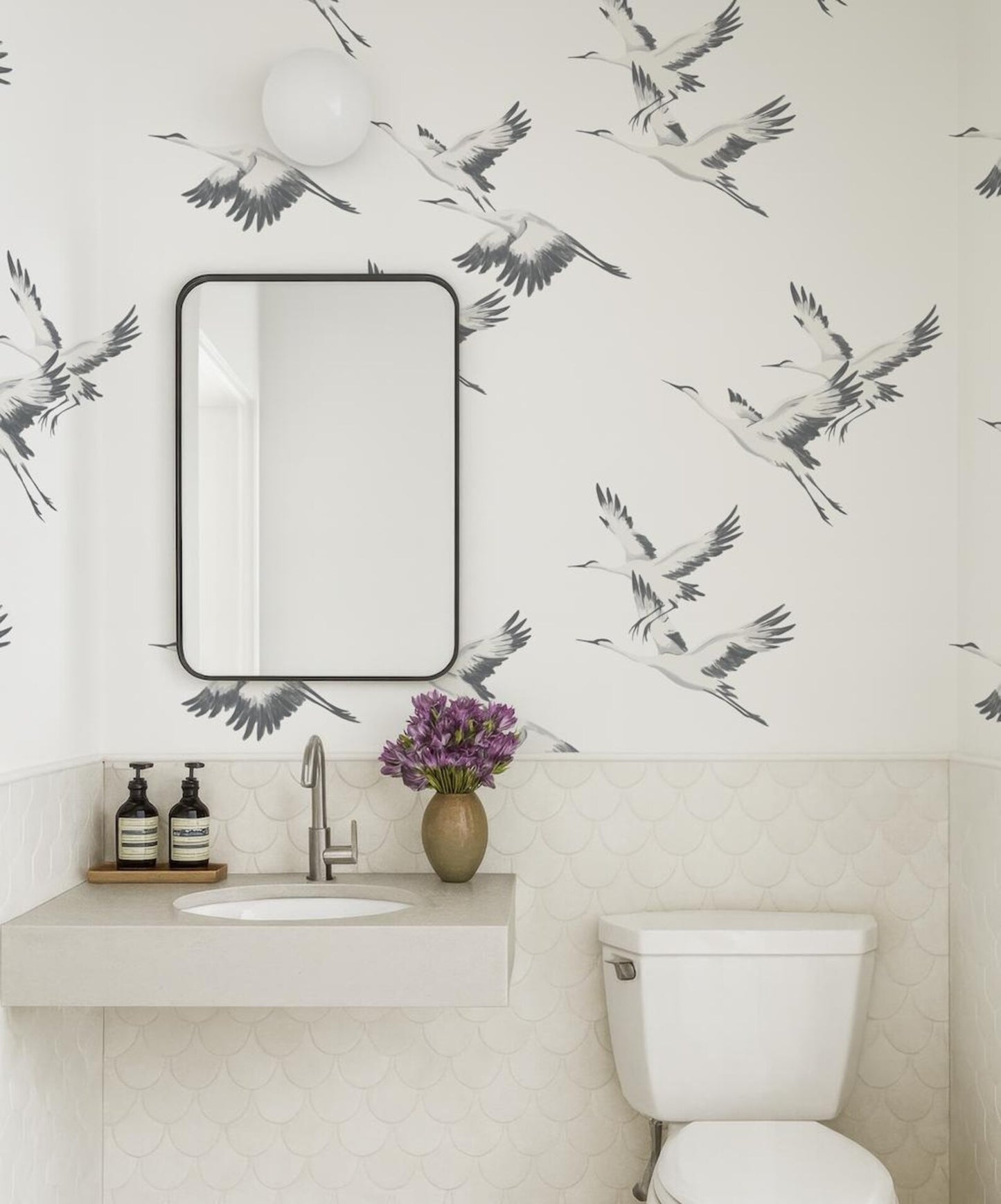 a bathroom with a white sink, a mirror, and a toilet. The walls are decorated with a black and white bird wallpaper, and there is a vase of purple flowers on the counter.
