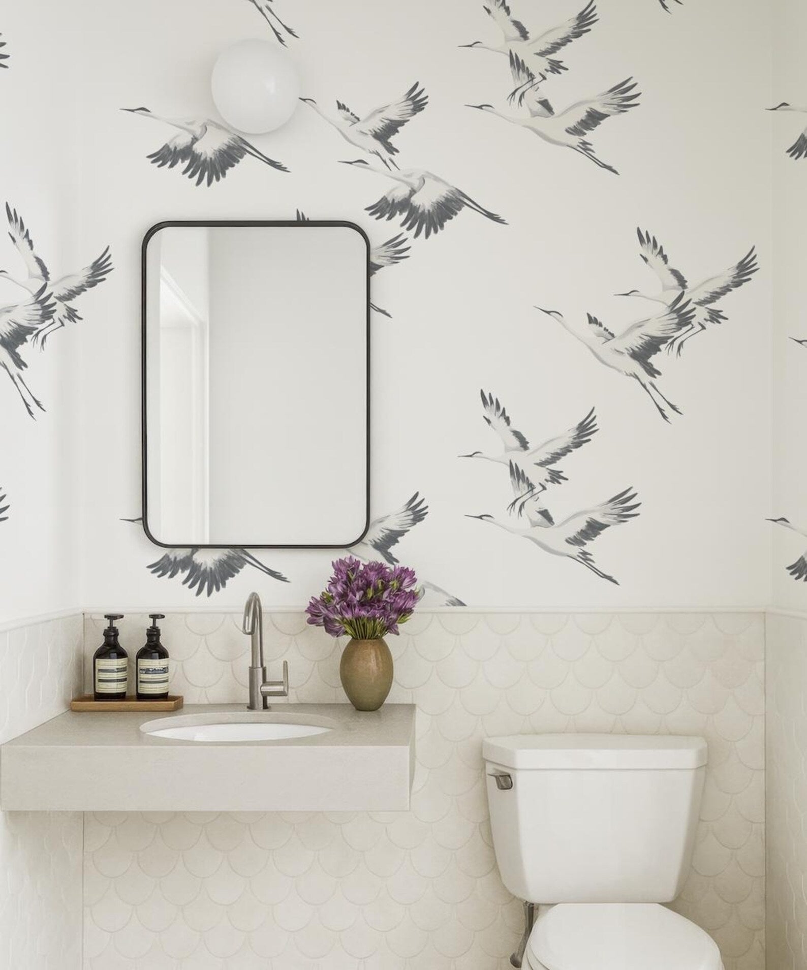 a bathroom with a white sink, a mirror, and a toilet. The walls are decorated with a black and white bird wallpaper, and there is a vase of purple flowers on the counter.