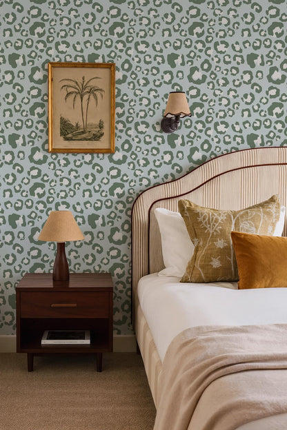 a bedroom with a bed, nightstand, and wall decor. The bed has a striped headboard and is adorned with throw pillows. The nightstand has a lamp and a framed picture on its surface. The wall behind the bed has a leopard print wallpaper, and there is a framed picture hanging above the bed.